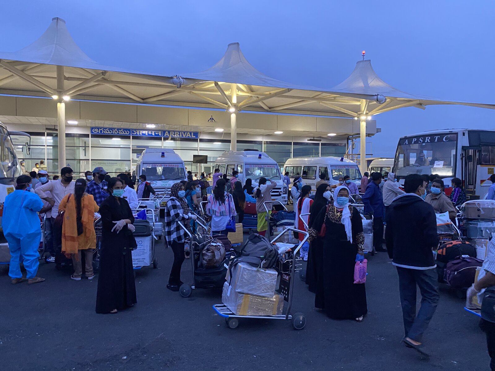 Crowd of travellers with luggage at Visakhapatnam Airport arrivals canopy at dusk · APSRTC bus and Vera Tempo Travellers + iMASQ buses lined up to process passengers