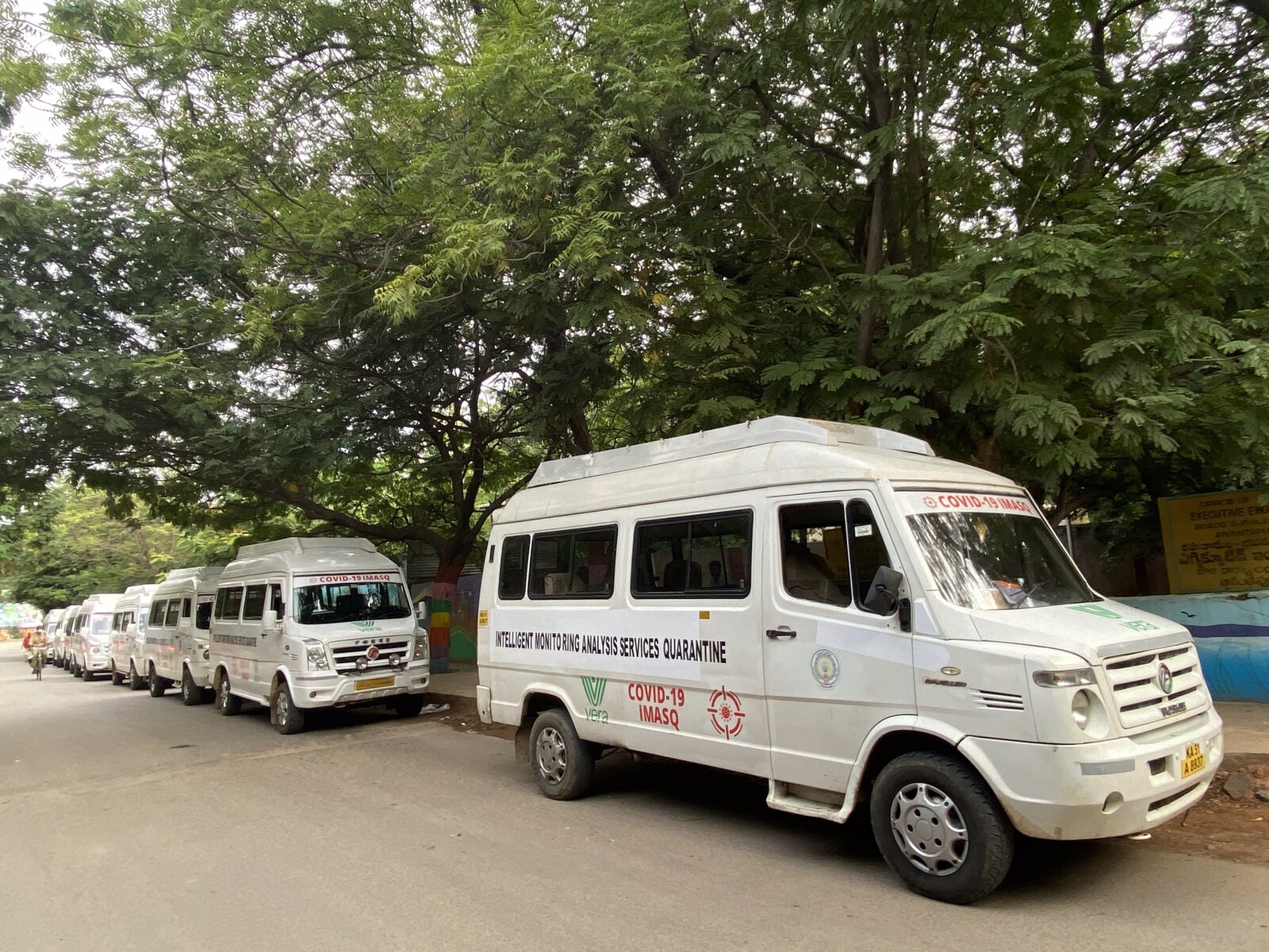 Vera iMASQ Tempo Traveller mini-bus fleet lined up in a parking area · INTELLIGENT MONITORING ANALYSIS SERVICES QUARANTINE branding · COVID-19 iMASQ red logo