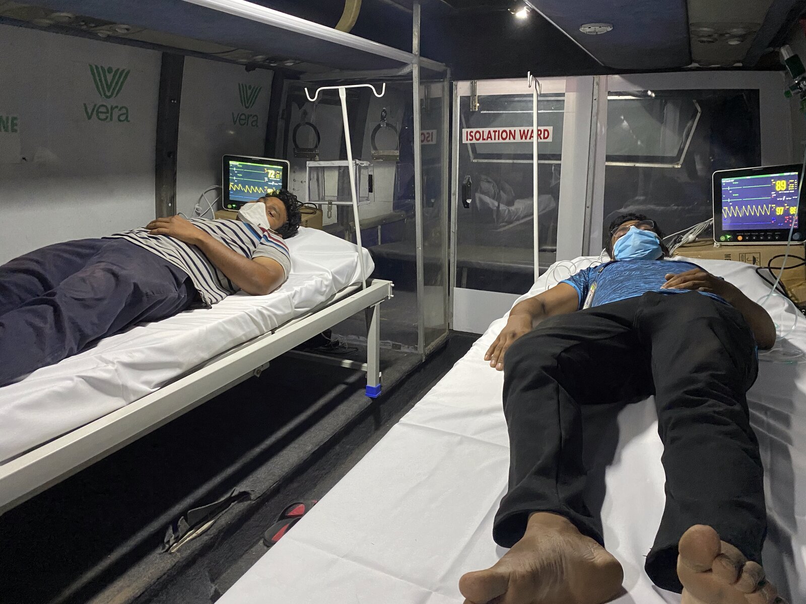 Two COVID patients in the b.SOZO Critical Care bus isolation ward · live ECG/vitals monitors showing real-time readings · Vera logo on the wall