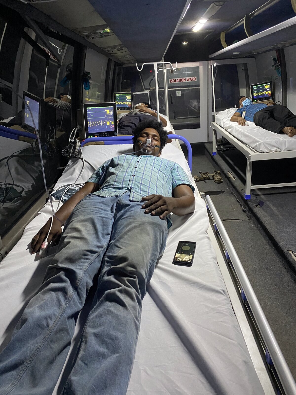 Patient on oxygen via nasal cannula inside the b.SOZO isolation ward · multiple additional patients visible on monitored beds in the background