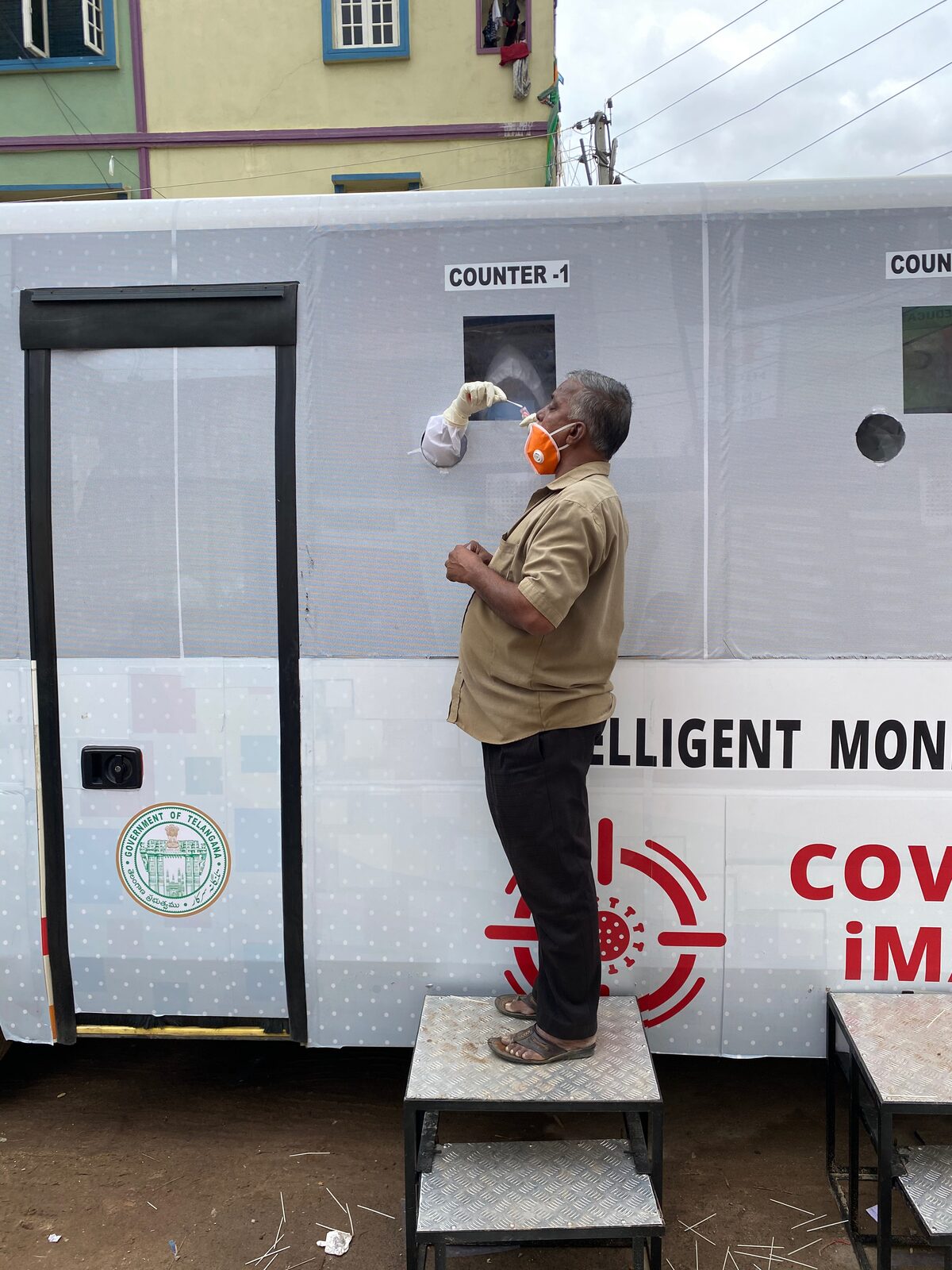 Citizen having nasal swab at Counter-1 of iMASQ bus · gloved hand reaching through the window with swab stick · Govt of Telangana seal visible · COVID-19 iMASQ red branding