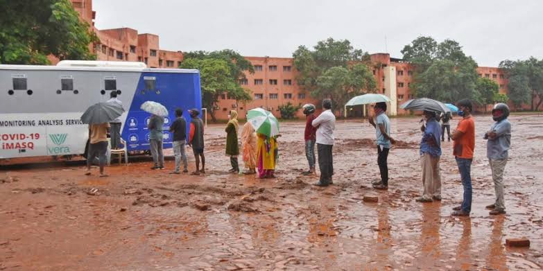 iMASQ blue bus deployed in heavy rain at outdoor site · people queued with umbrellas in red mud · Vera and COVID-19 iMASQ branding visible · crisis-grade deployment