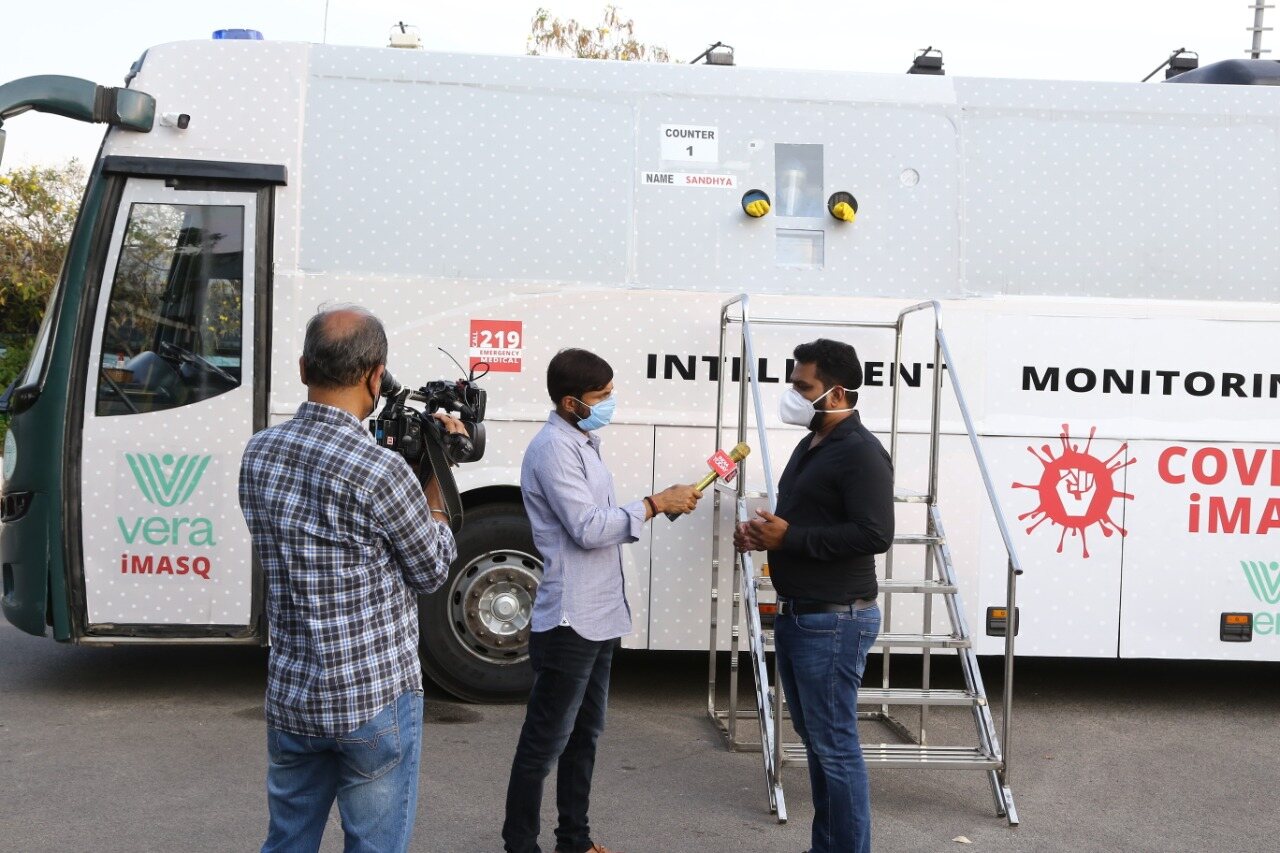 Dharma being interviewed by Aaj Tak / India Today outside the iMASQ bus · India Today microphone with red mic flag clearly visible · Vera iMASQ logo, COVID iMASQ red fist logo, and INTELLIGENT MONITORING branding all in frame · 108 Emergency Medical sticker on bus