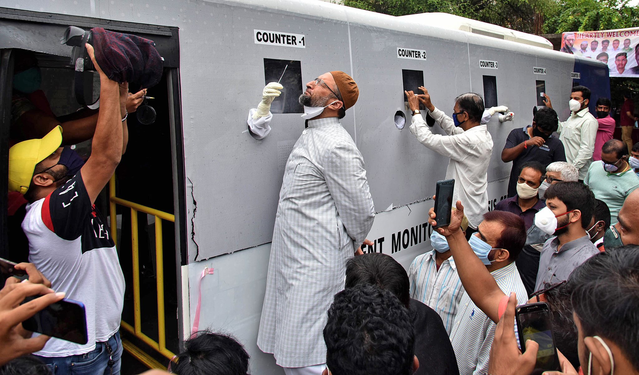 Asaduddin Owaisi, AIMIM President and Member of Parliament for Hyderabad, taking COVID nasal swab at iMASQ bus Counter-2 · INTELLIGENT MONITORING ANALYSIS SERVICES branding · Vera and iMASQ logos · welcoming banner
