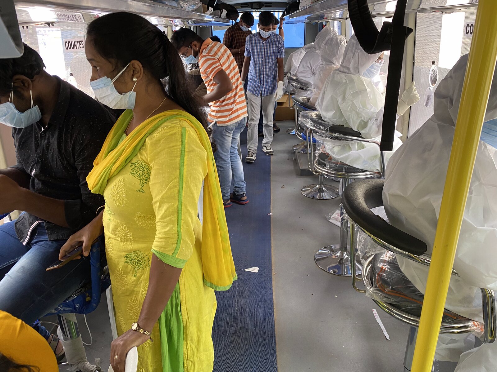 In-built Viral Transport Medium VTM packaging unit inside iMASQ bus · staff in green scrubs and PPE blue gown at sample-preparation desk · Vera branding
