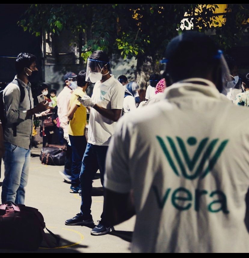 Vera-branded staff in white PPE T-shirts and face shields processing returning migrants at a transit hub · Vera logo visible on shirts